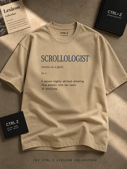 A beige graphic T-shirt laid flat on a textured surface with styled papers and a black box around it. The shirt has blue and black printed text reading 'SCROLLOLOGIST' with a dictionary-style definition underneath.