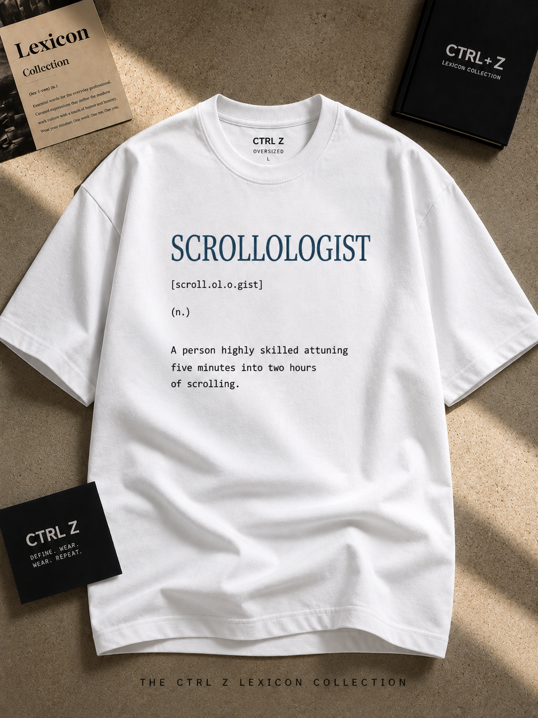 A white short-sleeve T-shirt laid flat on a textured beige surface, printed with the word 'SCROLLOLOGIST' and a dictionary-style definition in dark blue and black text. A black box and branded card labeled CTRL Z are visible around it, suggesting branded packaging or promotional materials.